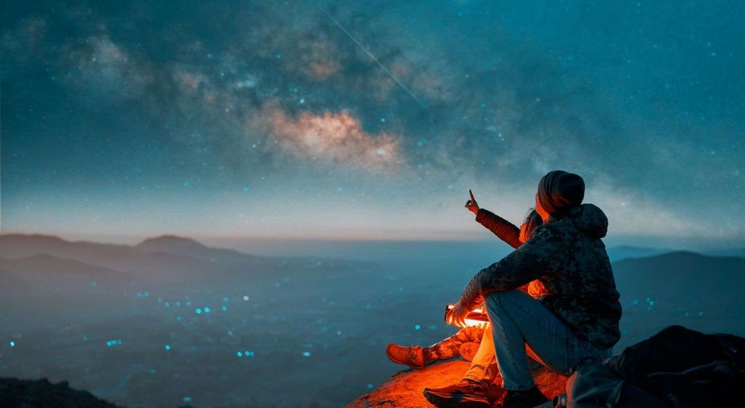 Silhouettes of a couple sitting on the top of the hill looking at shooting stars and the milky way in the background