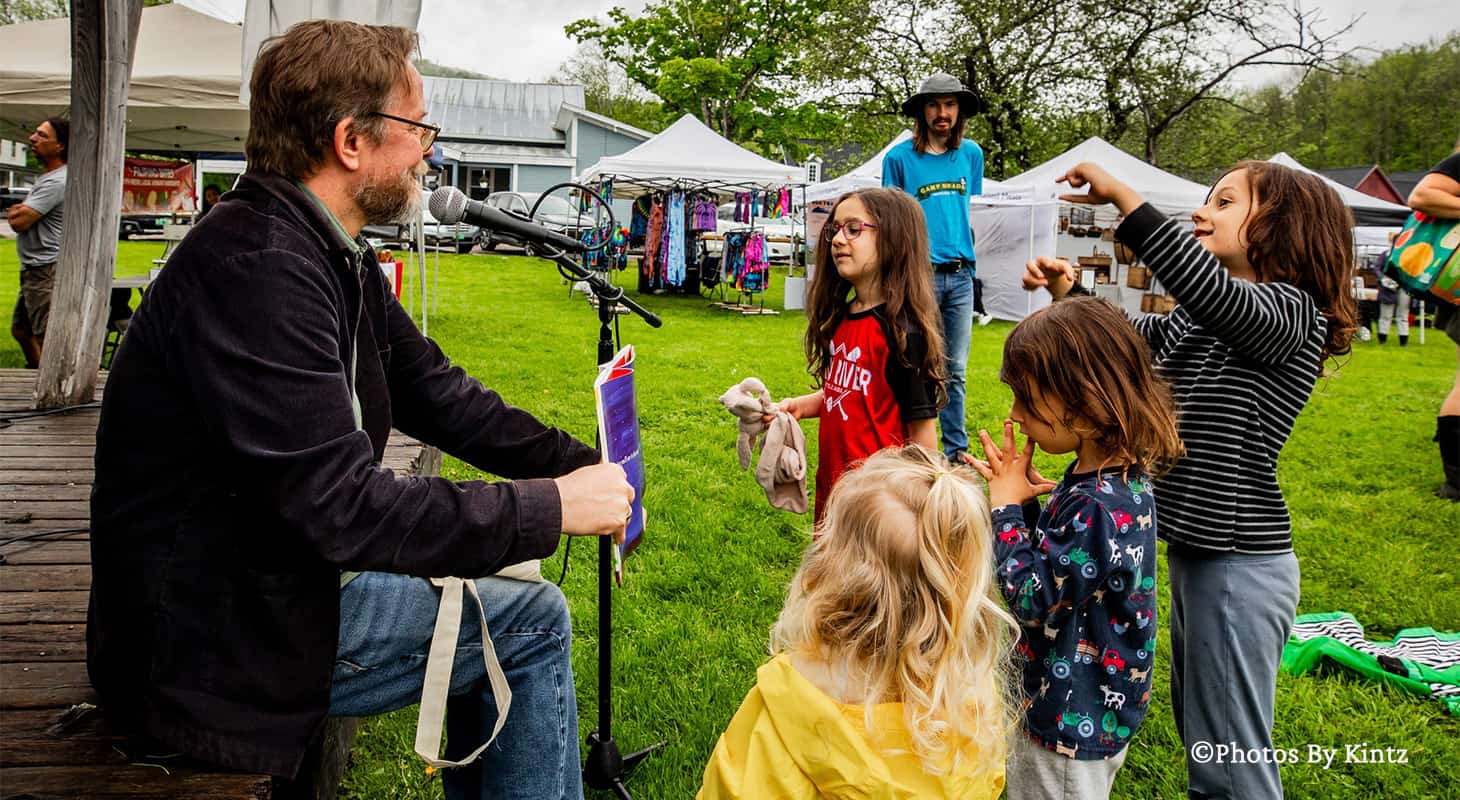 A man reading to a group of kids on the stage at a Vermont Farmers Market in Waitsfield