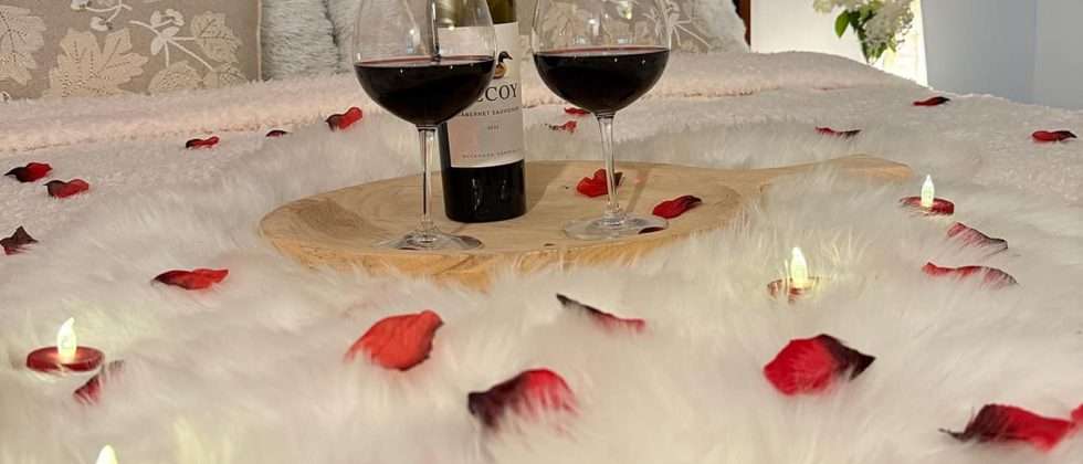 Two wine glasses, bottle of wine, rose petals, and tea lights on a bed